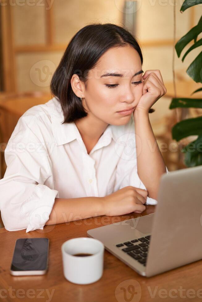 Vertical shot of asian girl looking tired at laptop screen, bored of working, sitting in cafe sleepy and exhausted photo