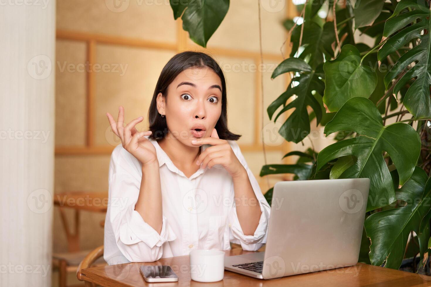 Girl with surprised face, pitching an idea, pointing left, sitting with laptop in cafe and drinking coffee photo