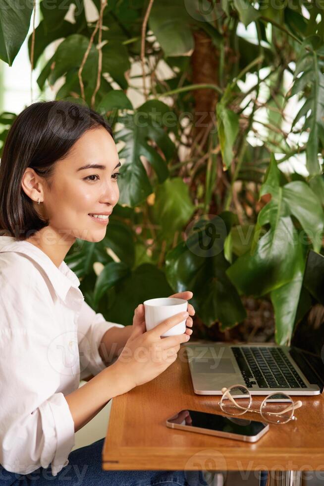 Vertical shot of beautiful woman sits in cafe with laptop, works or studies online at co-working space, smiling relaxed photo
