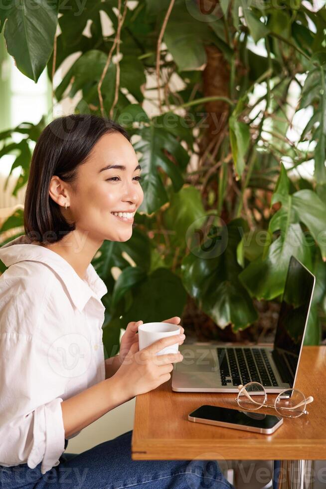 Vertical shot of beautiful woman sits in cafe with laptop, works or studies online at co-working space, smiling relaxed photo