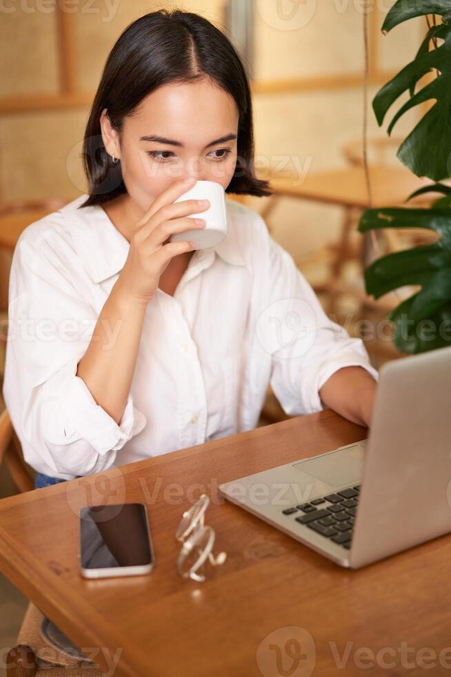 Student sitting in cafe with cup of coffee. Young asian woman working on laptop in restaurant, sitting with computer and smartphone photo