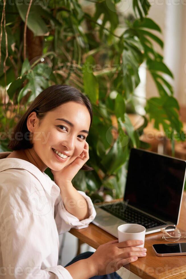 Trendy young woman sitting in cafe and smiling at camera, drinking coffee and using laptop, working remotely, studying or browsing internet photo