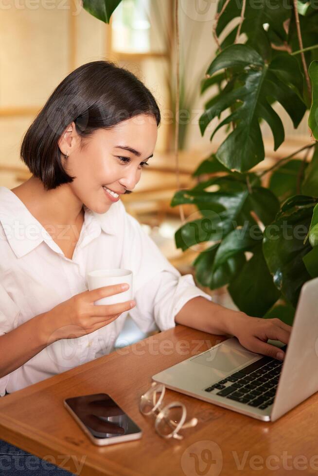 Student sitting in cafe with cup of coffee. Young asian woman working on laptop in restaurant, sitting with computer and smartphone photo