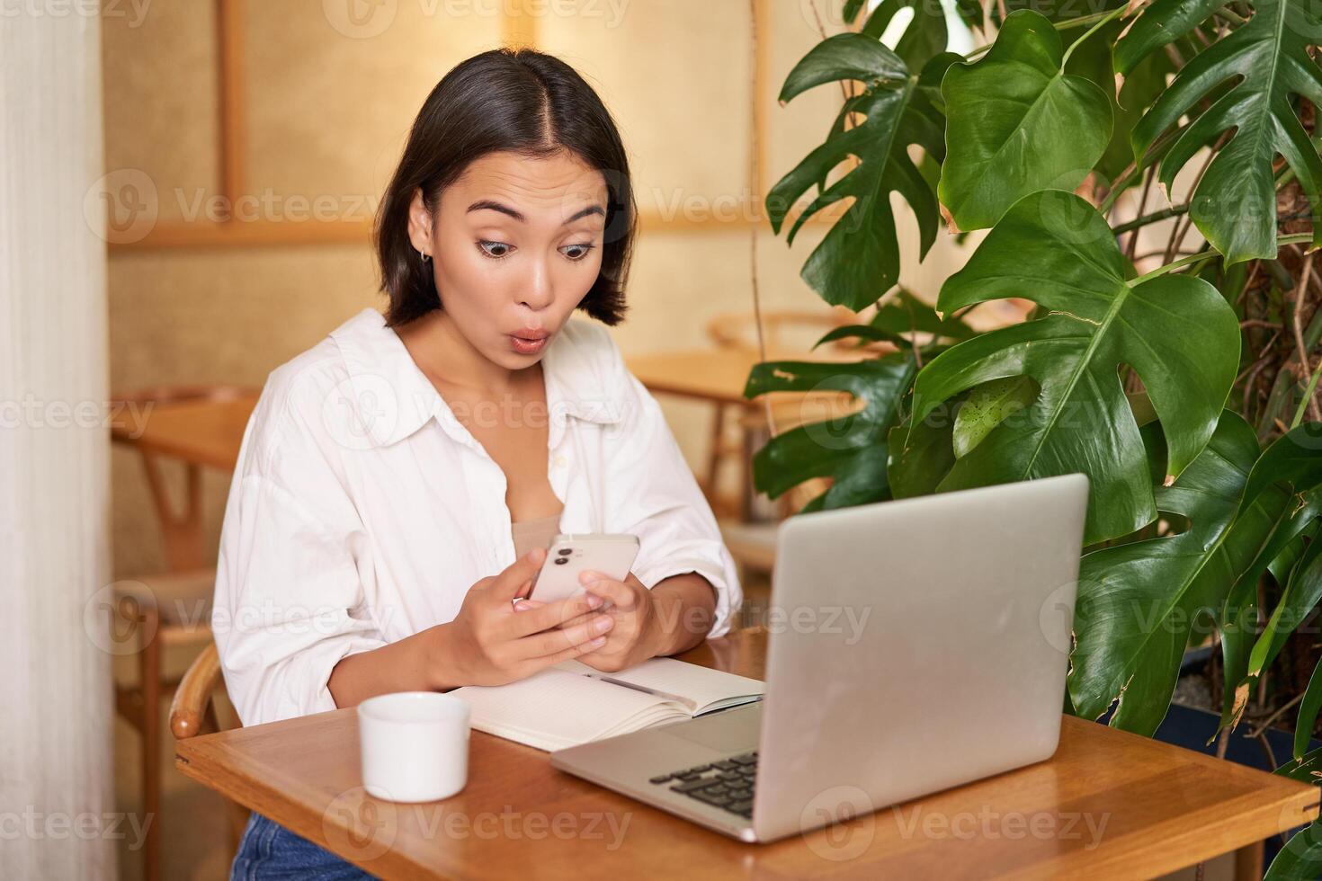 Woman in cafe sitting with laptop, looking surprised at smartphone screen, reading exciting good news photo