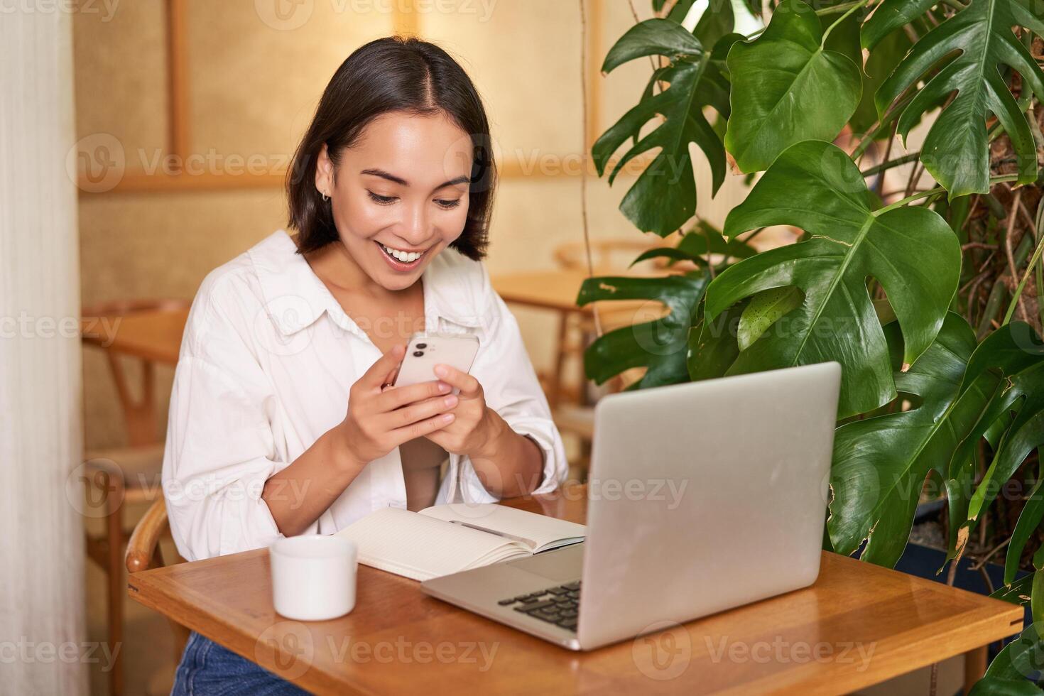 Woman in cafe sitting with laptop, looking surprised at smartphone screen, reading exciting good news photo