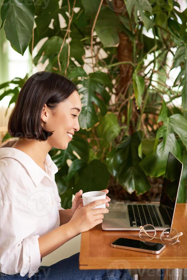 Vertical shot of beautiful woman sits in cafe with laptop, works or studies online at co-working space, smiling relaxed photo