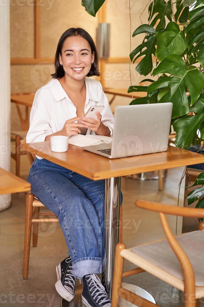 Vertical shot of young asian girl sits in cafe with laptop and smartphone, relaxing and surfing the net, working remotely photo