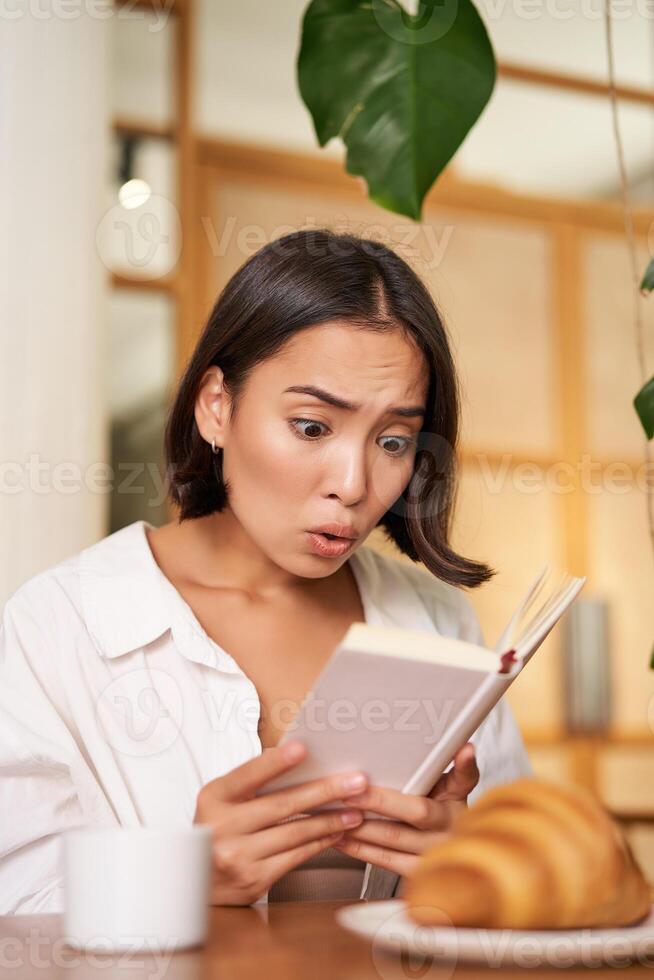 Vertical shot of asian girl looking shocked and worried at book, reading with interest, gasping from story plot, sitting in cafe with croissant and coffee photo