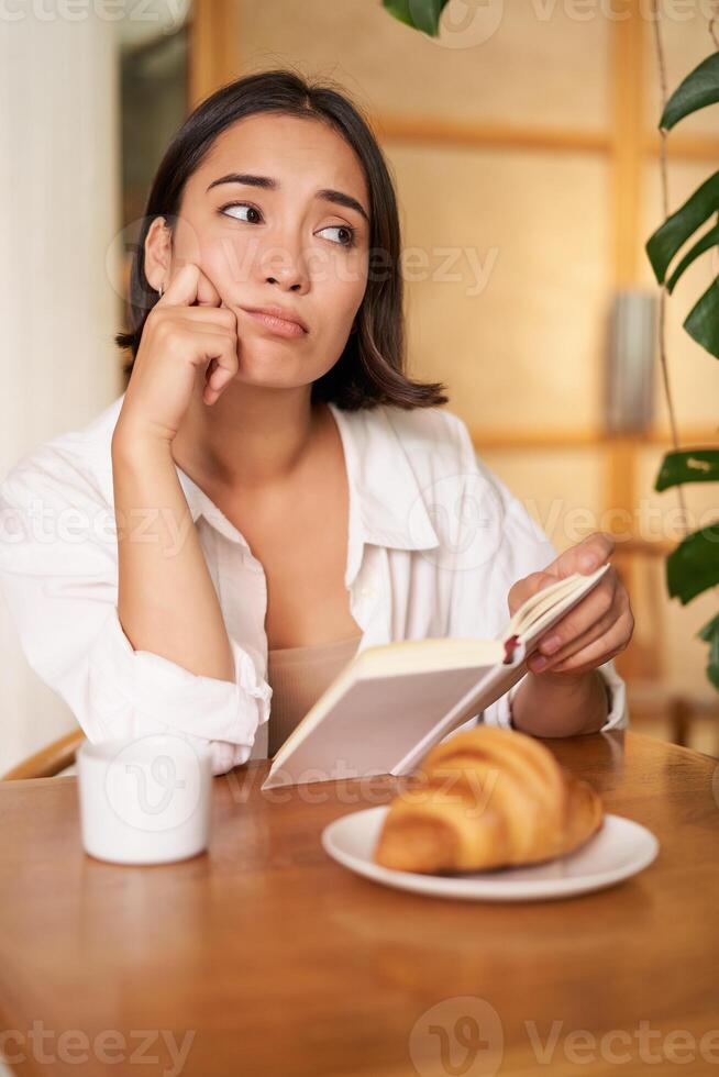 Young woman feels uneasy, reads sad book and sits in cafe with cup of coffee, looks around with sulking unhappy face photo