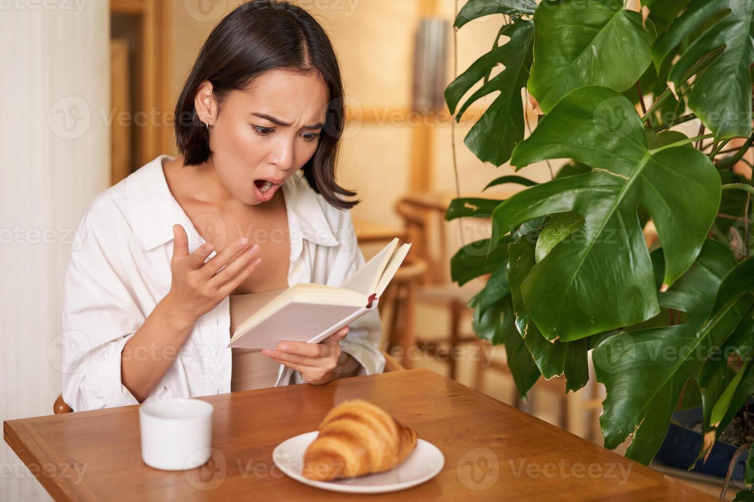 Portrait of asian woman looking shocked at book pages, reading something interesting, concentrating, sitting in cafe with coffee and croissant photo
