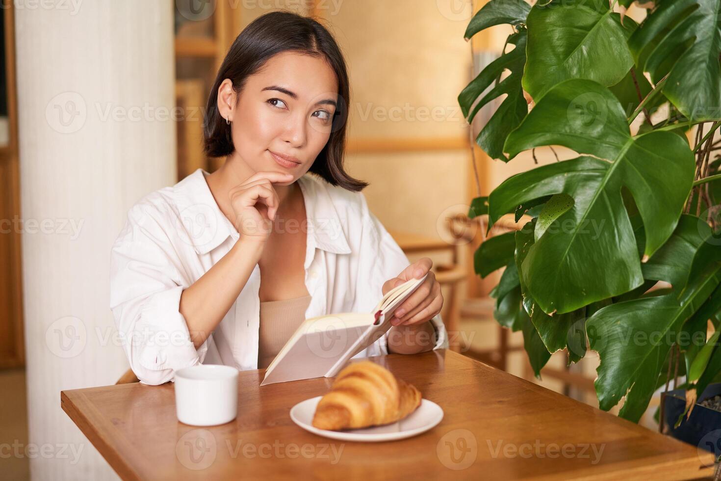 Smiling beautiful girl with a book, reading with thoughtful smirk, thinking of something, drinking coffee in cafe photo