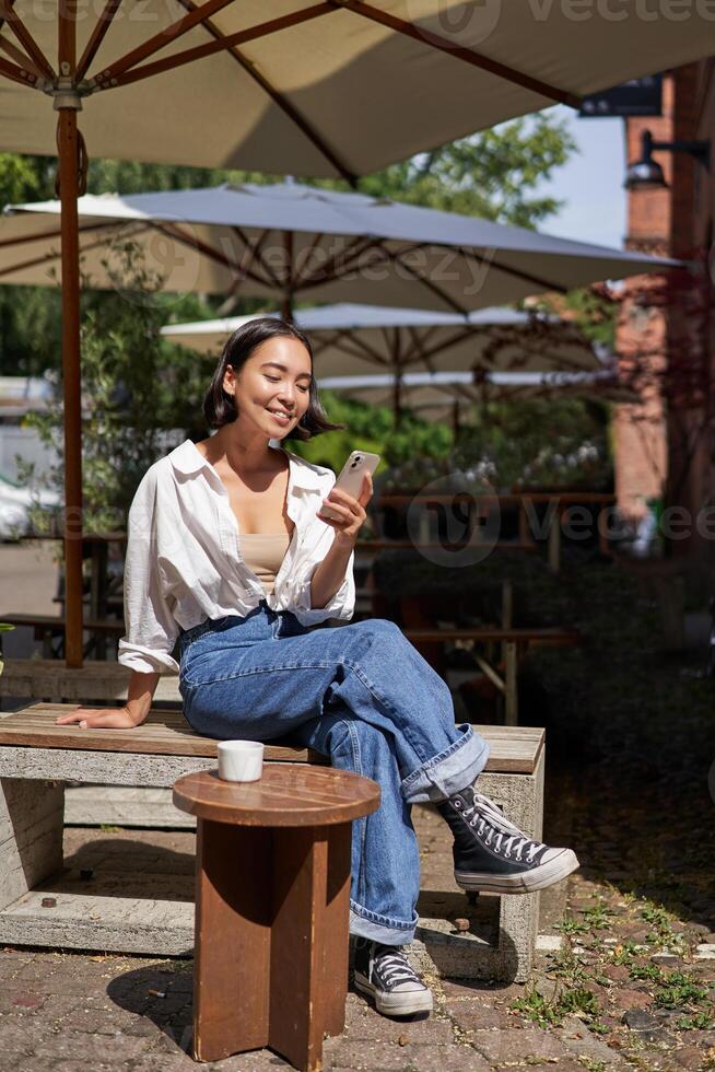 People and communication. Stylish asian girl sits in outdoor cafe with cup of coffee and smartphone, using mobile phone photo