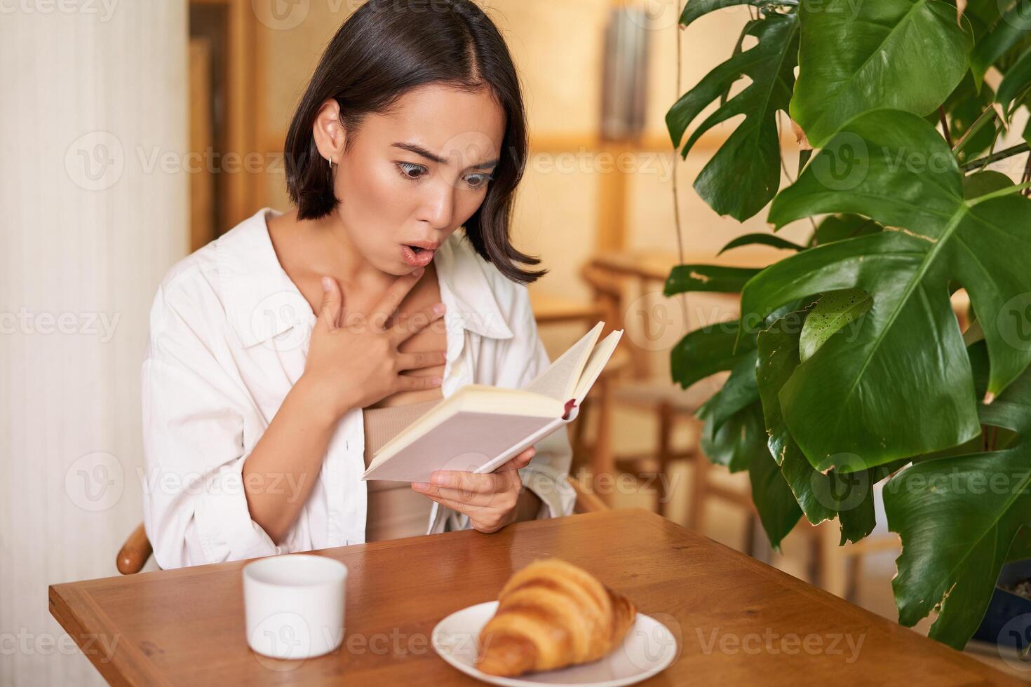 Portrait of asian woman looking shocked at book pages, reading something interesting, concentrating, sitting in cafe with coffee and croissant photo