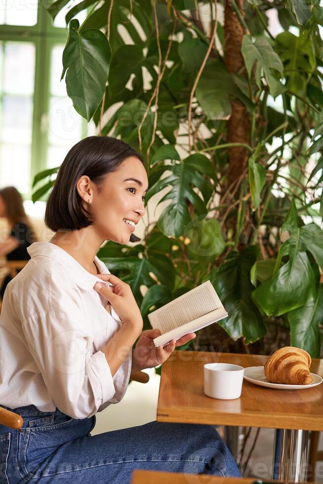 Vertical shot of asian brunette girl sitting elegant with book, reading in cafe and smiling, drinking coffee photo