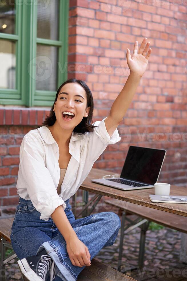 Vertical shot of happy girl attracting attention, waving hand at friend in cafe, sitting on bench with laptop photo