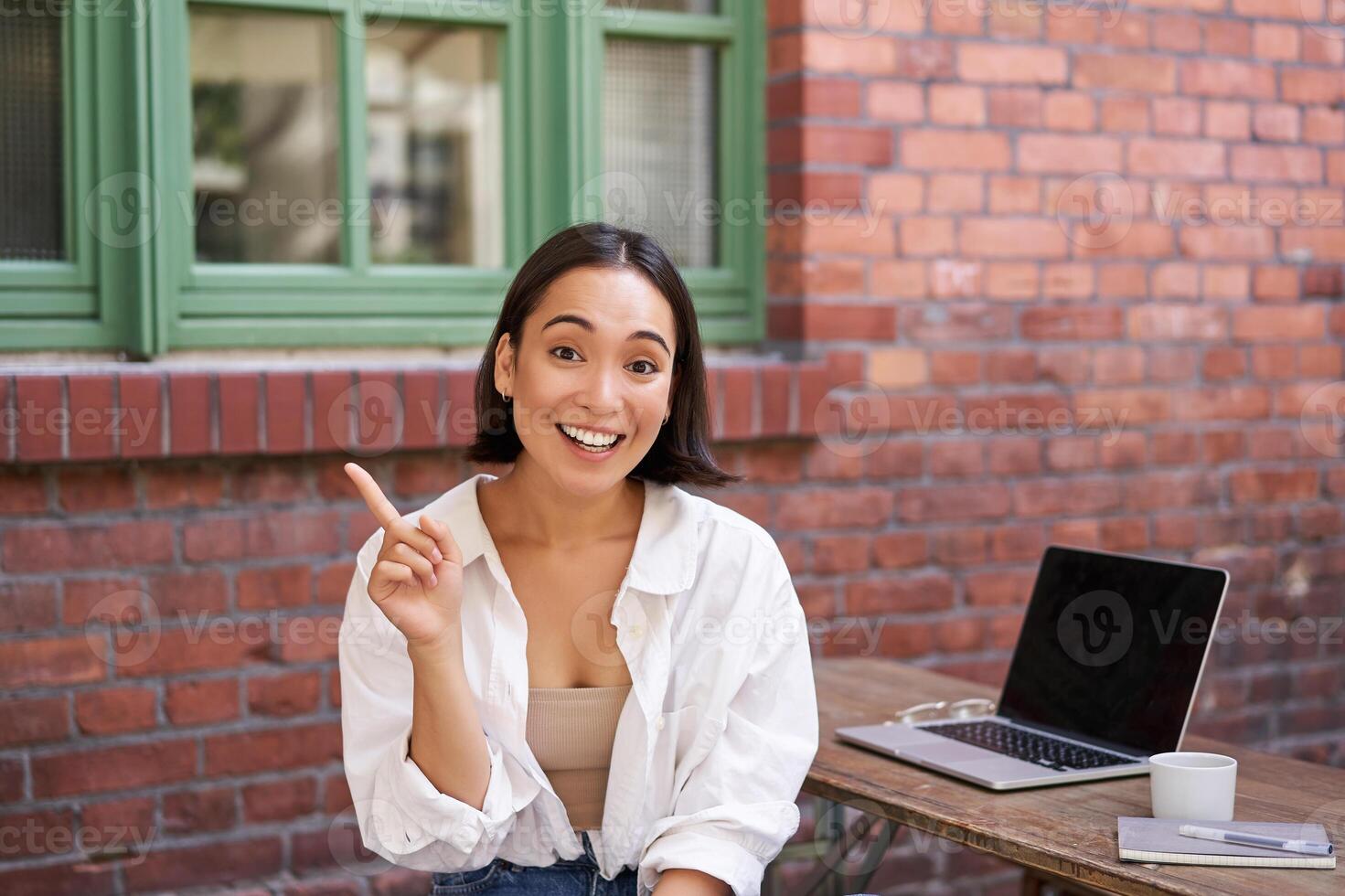 Asian girl with surprised face, sitting with laptop, pointing left at copy space, banner with information, looking amazed photo