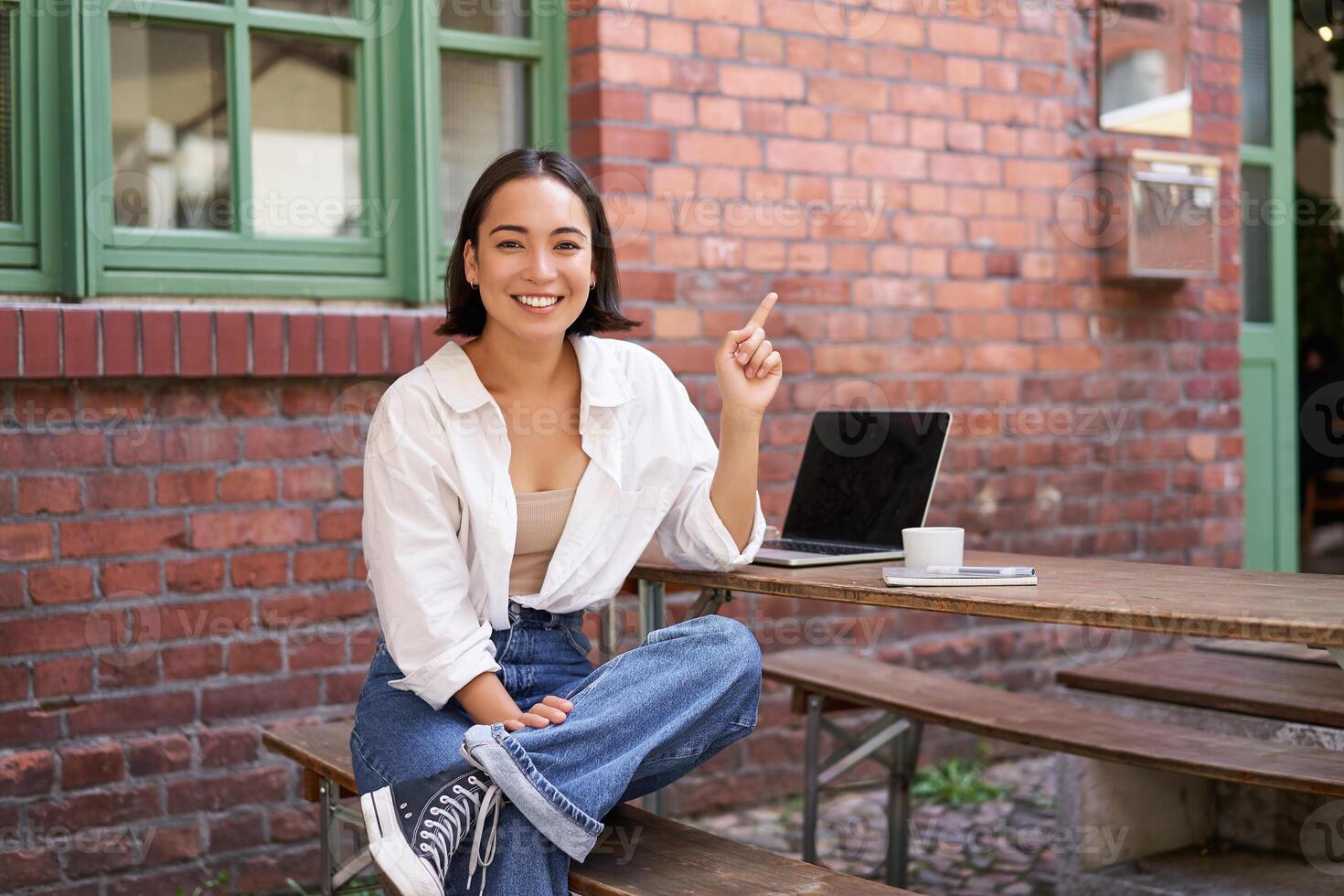 stylish modern asian girl with laptop, sitting in cafe, looking amazed and pointing at upper right corner banner, showing info advertisement photo