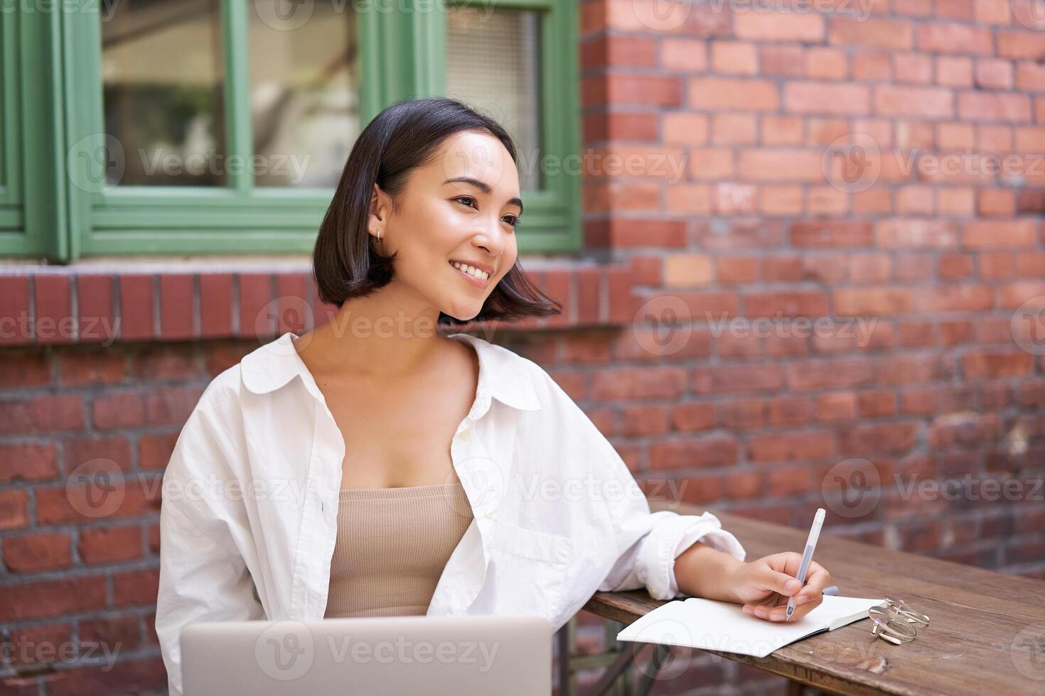 Beautiful young female model sitting with laptop in outdoor cafe, taking notes, writing in her diary or working on project, doing homework photo