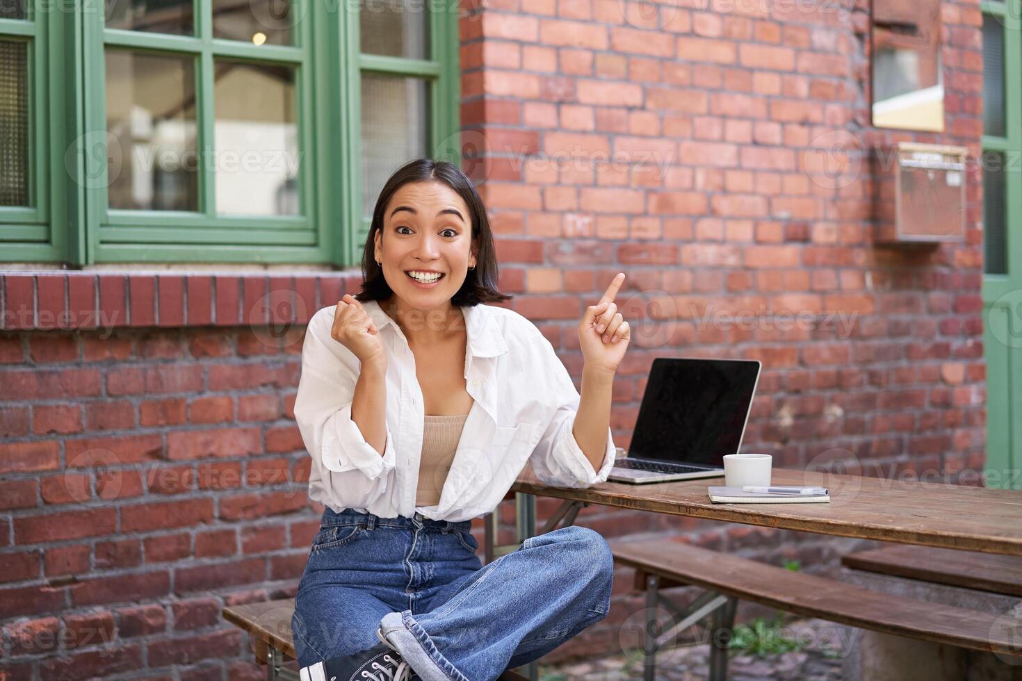 Stylish urban girl, asian woman with laptop sits in cafe, points at upper right corner banner, shows copy space with surprised face expression photo