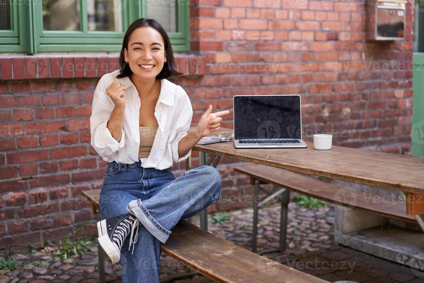Happy korean girl with laptop, sitting in cafe, laughing and pointing at copy space on brick wall, showing advertisement photo