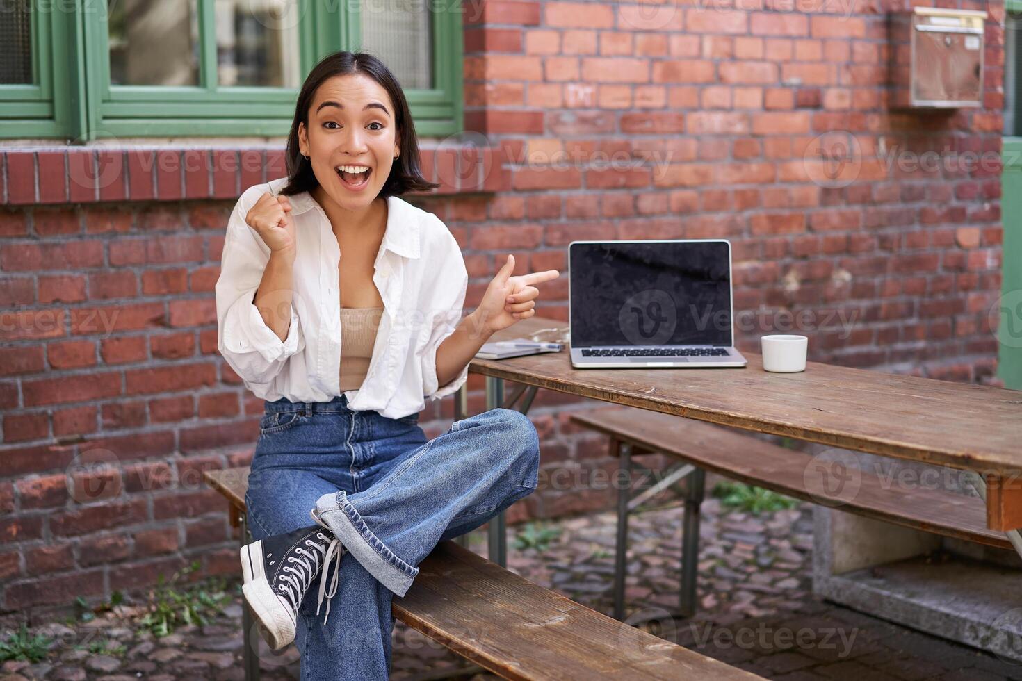 Happy korean girl with laptop, sitting in cafe, laughing and pointing at copy space on brick wall, showing advertisement photo