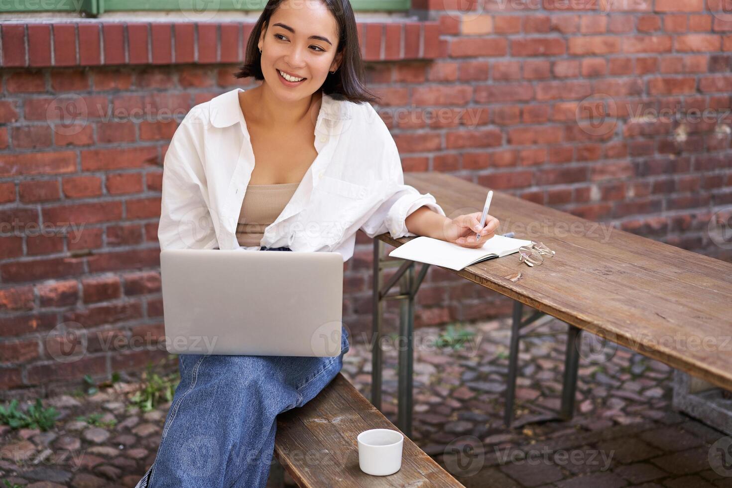 Working woman sitting outdoors in cafe, looking at laptop and making notes in notebook, writing down while watching smth on computer photo