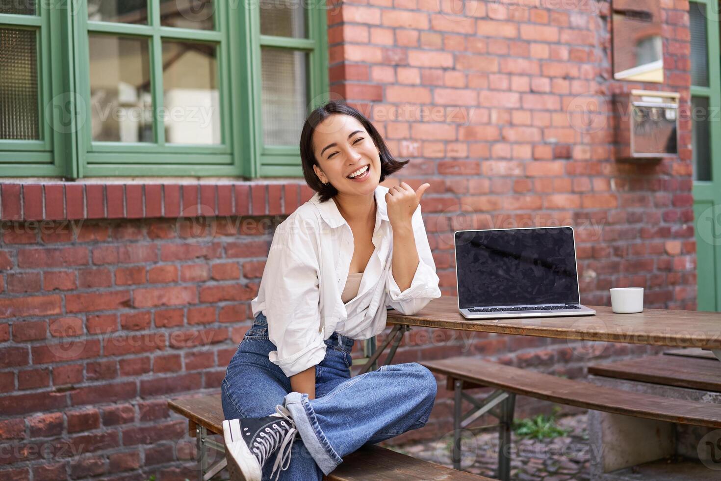 Happy korean girl with laptop, sitting in cafe, laughing and pointing at copy space on brick wall, showing advertisement photo