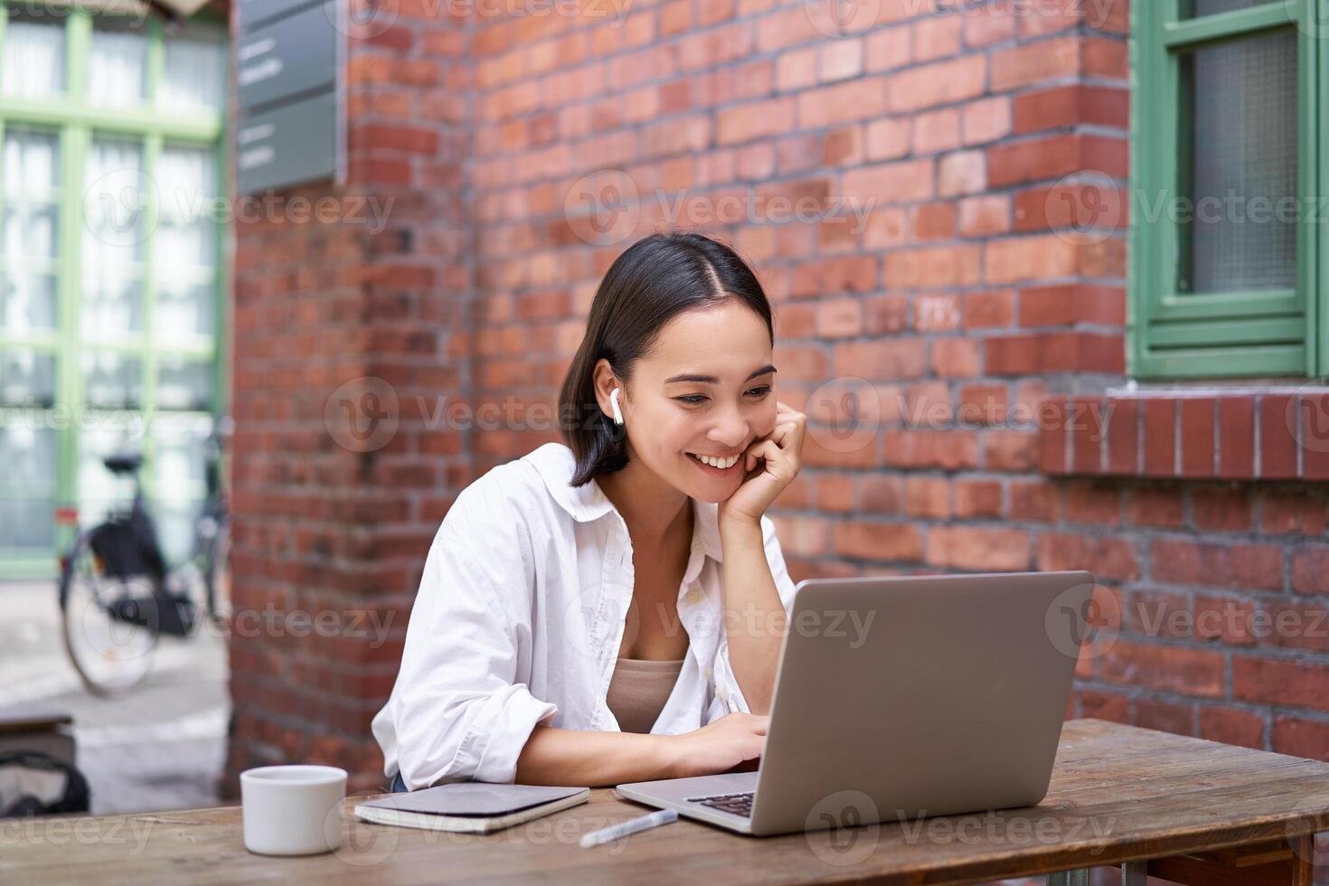 Working woman sitting in coworking space, drinking coffee and using laptop, wearing wireless headphones, watching video and smiling photo