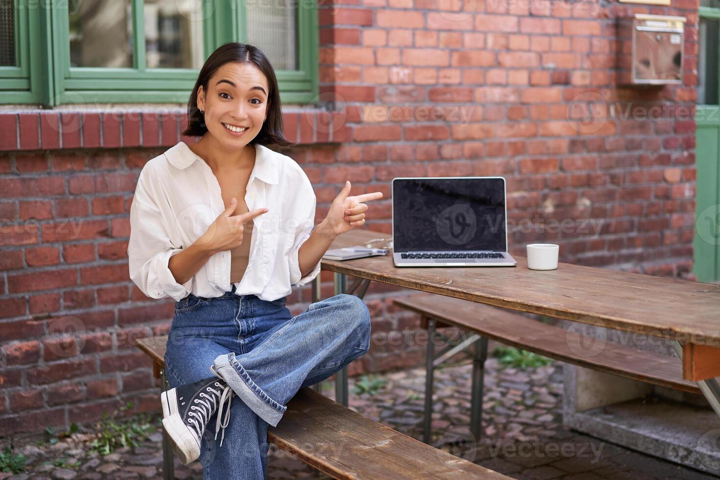 Stylish young asian woman sitting in cafe with laptop, outdoor table, pointing finger at advertisement, showing banner copy space photo