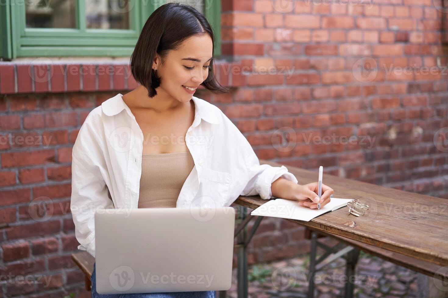 Working woman sitting outdoors in cafe, looking at laptop and making notes in notebook, writing down while watching smth on computer photo