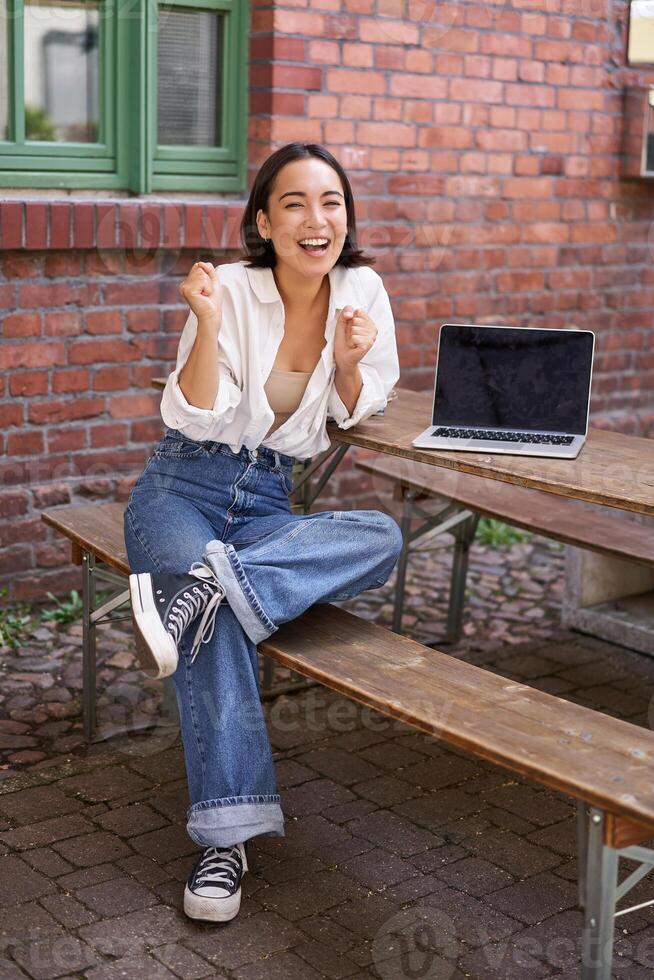 Vertical shot of enthusiastic asian girl, sits with laptop, screen is empty and blank, woman laughing and celebrating, triumphing photo
