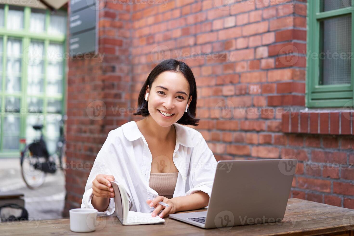 Working woman sitting in coworking space, drinking coffee and using laptop, wearing wireless headphones, watching video and smiling photo