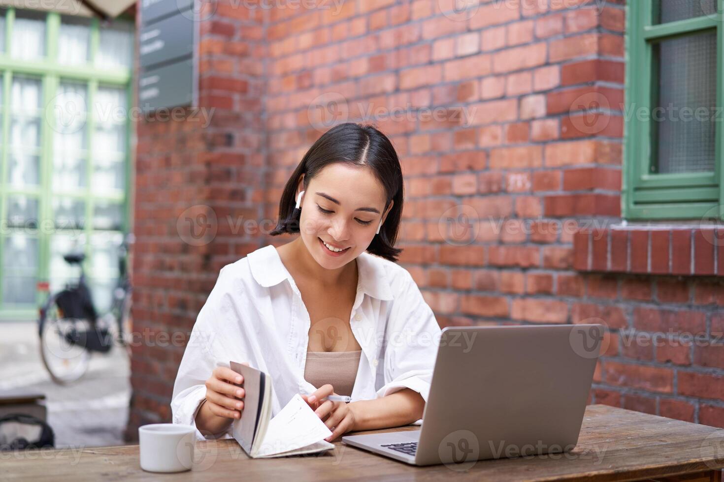 Working woman sitting in coworking space, drinking coffee and using laptop, wearing wireless headphones, watching video and smiling photo