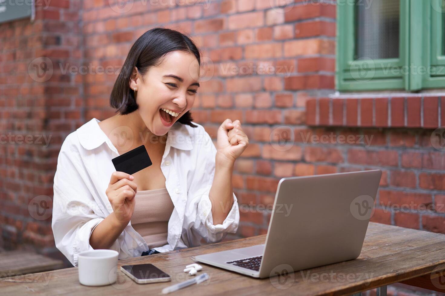 Happy young asian woman sitting near laptop, holding credit card, paying bills, shopping online contactless, smiling at camera photo