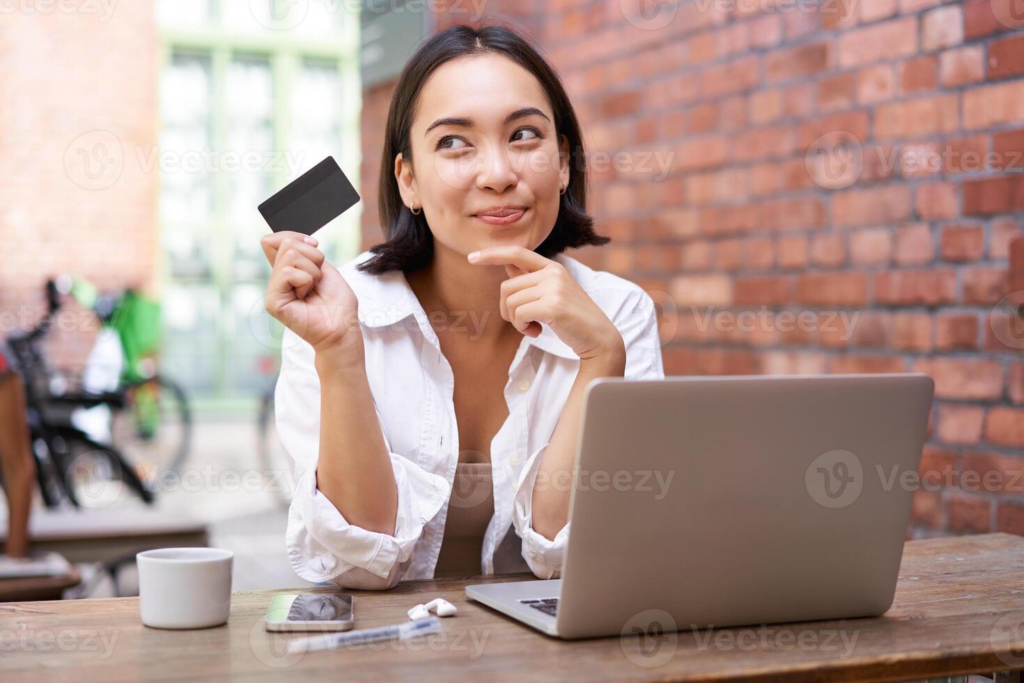Stylish young asian woman doing shopping online, sitting with credit card and laptop, buying in internet and paying contactless, dealing with bills photo