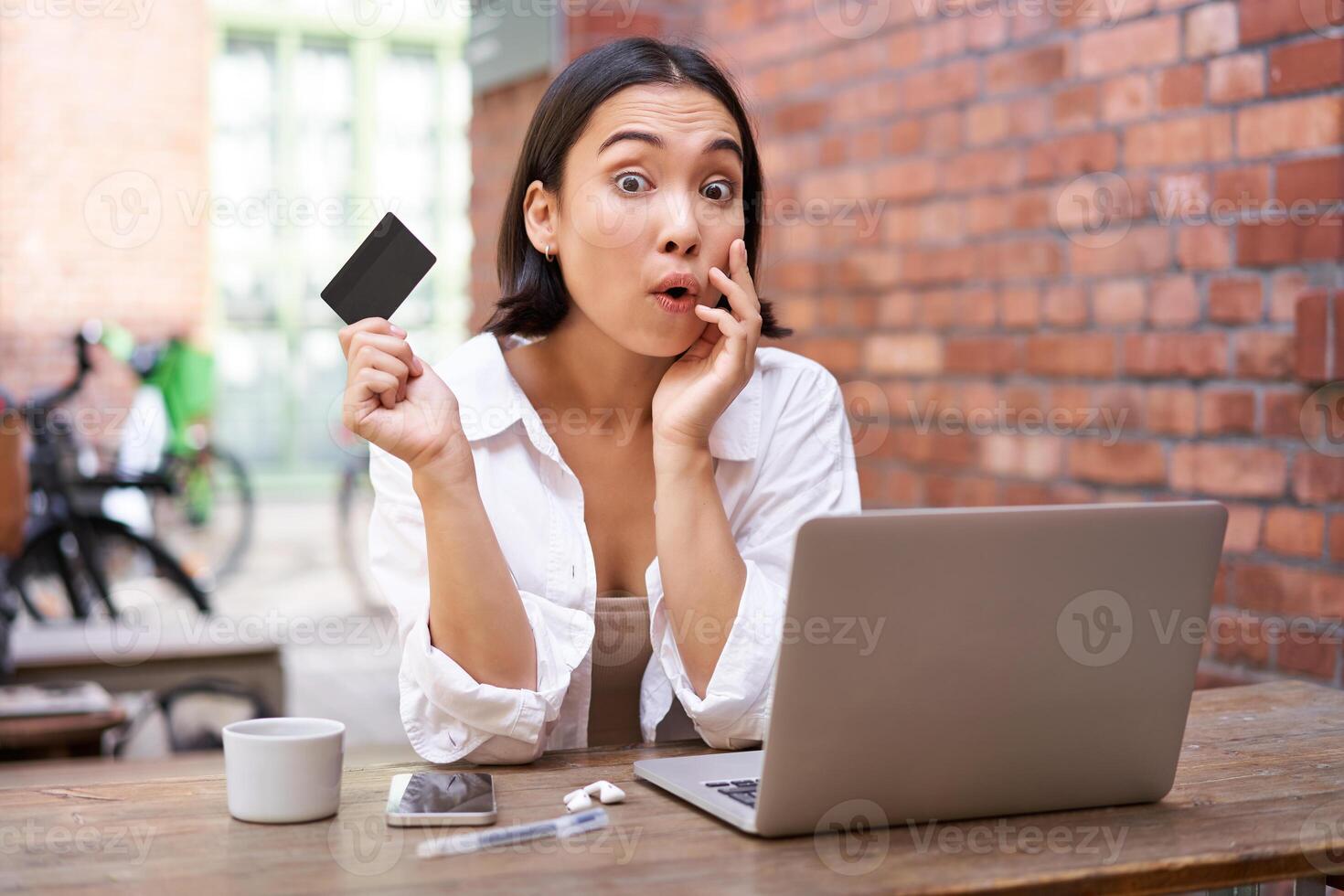 Stylish young asian woman doing shopping online, sitting with credit card and laptop, buying in internet and paying contactless, dealing with bills photo