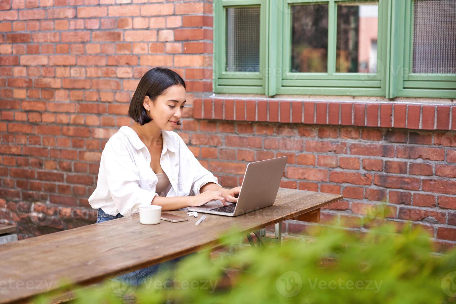 Stylish young urban girl in outdoor cafe, sitting on bench with laptop, smiling, browing on computer photo