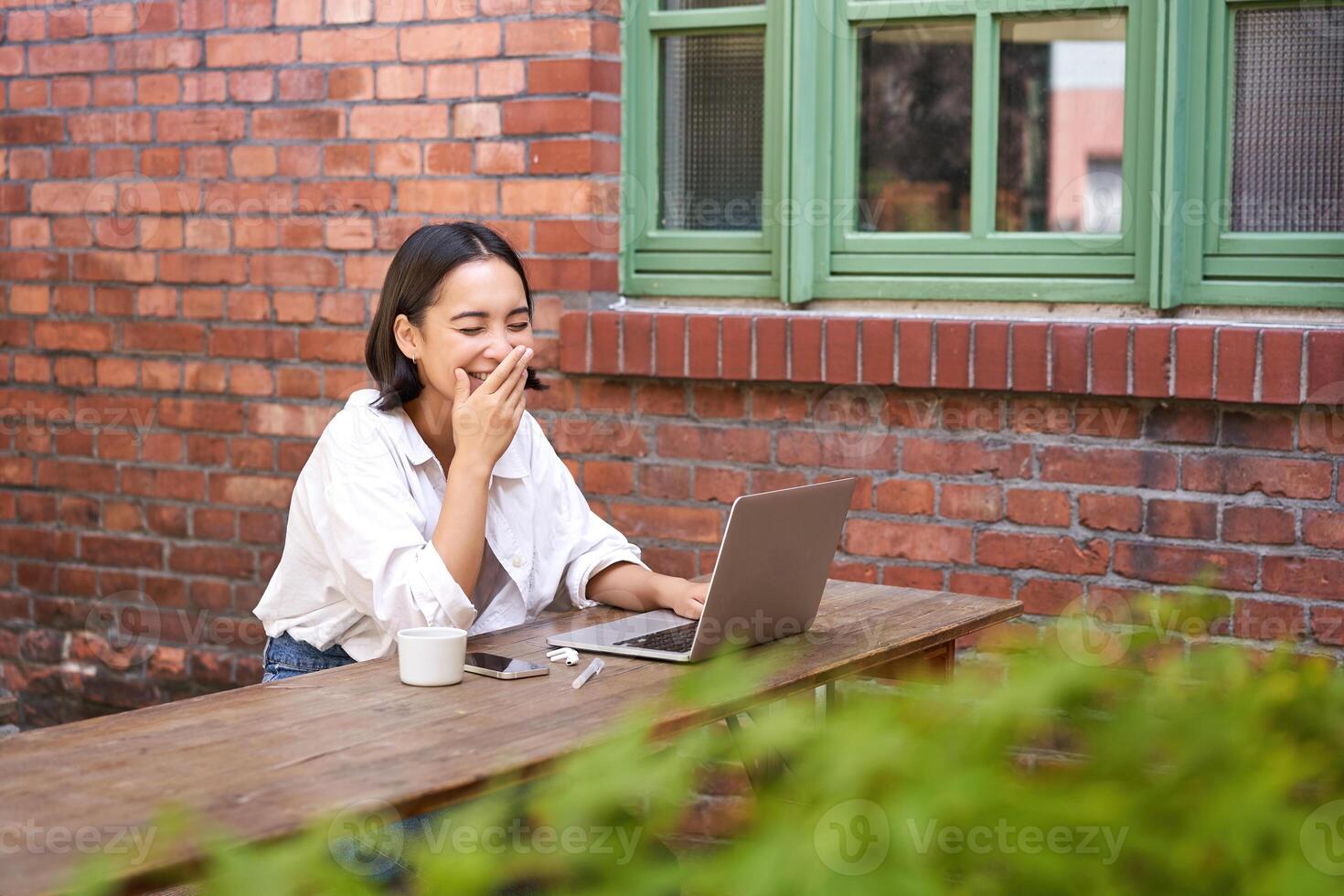 Stylish young urban girl in outdoor cafe, sitting on bench with laptop, smiling, browing on computer photo