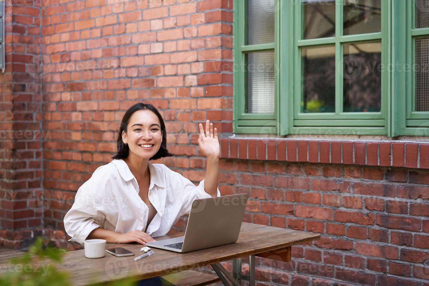 Friendly asian woman sitting with laptop, waving at you, saying hello, hi gesture, greeting you while working with computer photo
