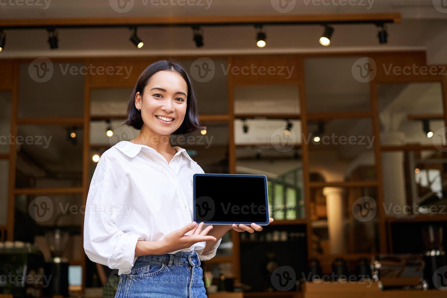 Smiling asian woman showing digital tablet screen, cafe owner showing smth, standing in front of cafe entrance photo