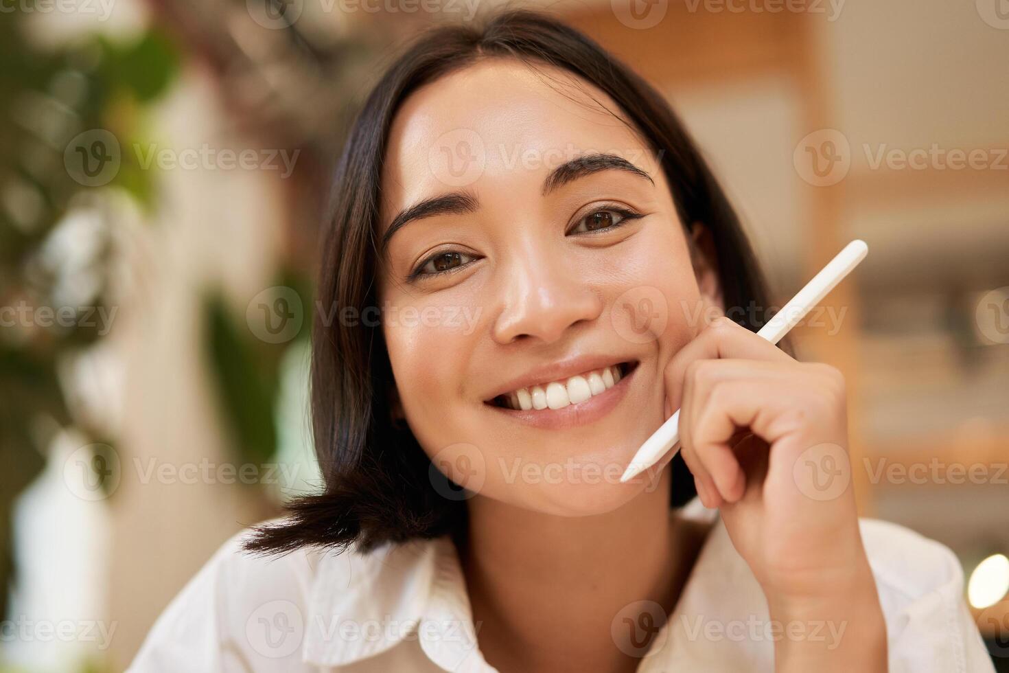 Close up portrait of smiling brunette asian woman, sitting with graphic pen and looking happy, drawing on digital tablet photo