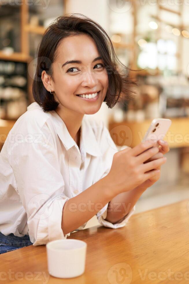 Vertical shot of brunette asian girl sitting in cafe, using smartphone app, chatting on mobile phone and smiling photo