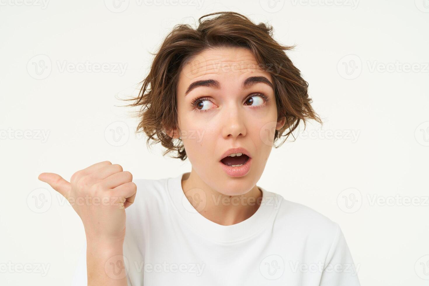 Image of girl with surprised face, points finger left, looks shocked, poses over white studio background photo