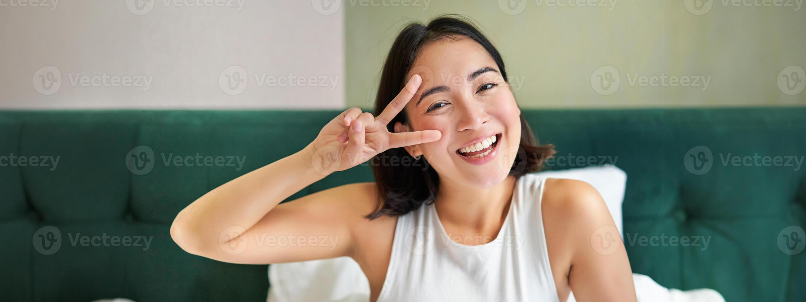 Positive asian woman lying in bed, showing peace sign, enjoys happy morning, waking up upbeat, staying in her bedroom photo