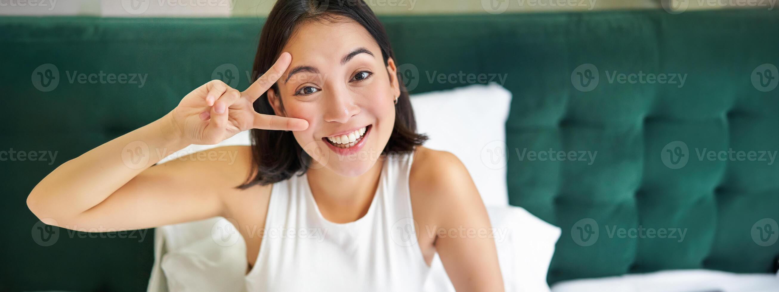 Positive asian woman lying in bed, showing peace sign, enjoys happy morning, waking up upbeat, staying in her bedroom photo