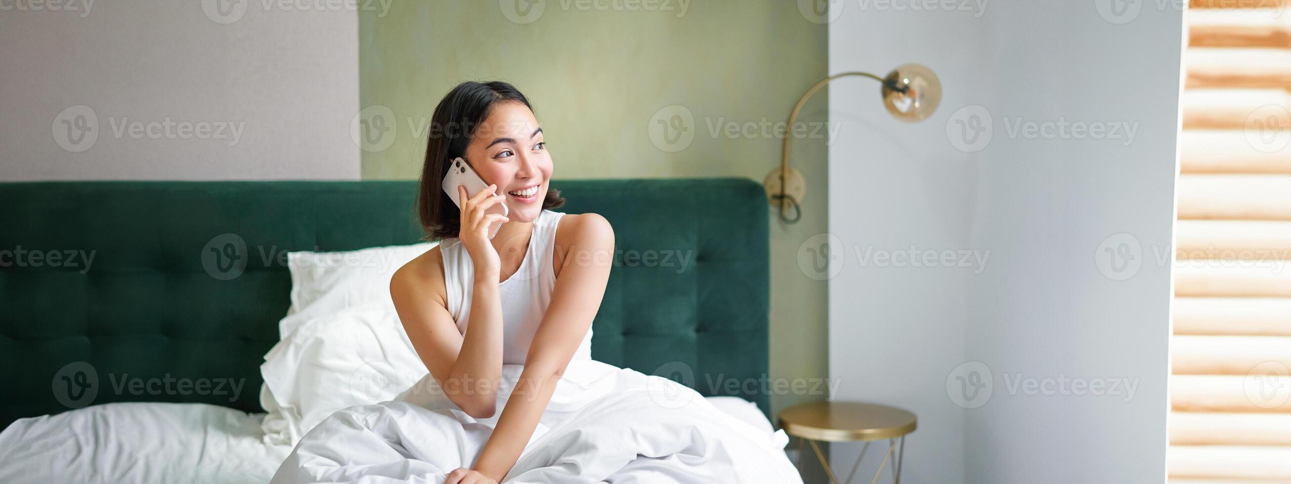 Beautiful smiling asian girl talking on mobile phone, lying in bed with pleased happy face, speaking to someone on telephone photo