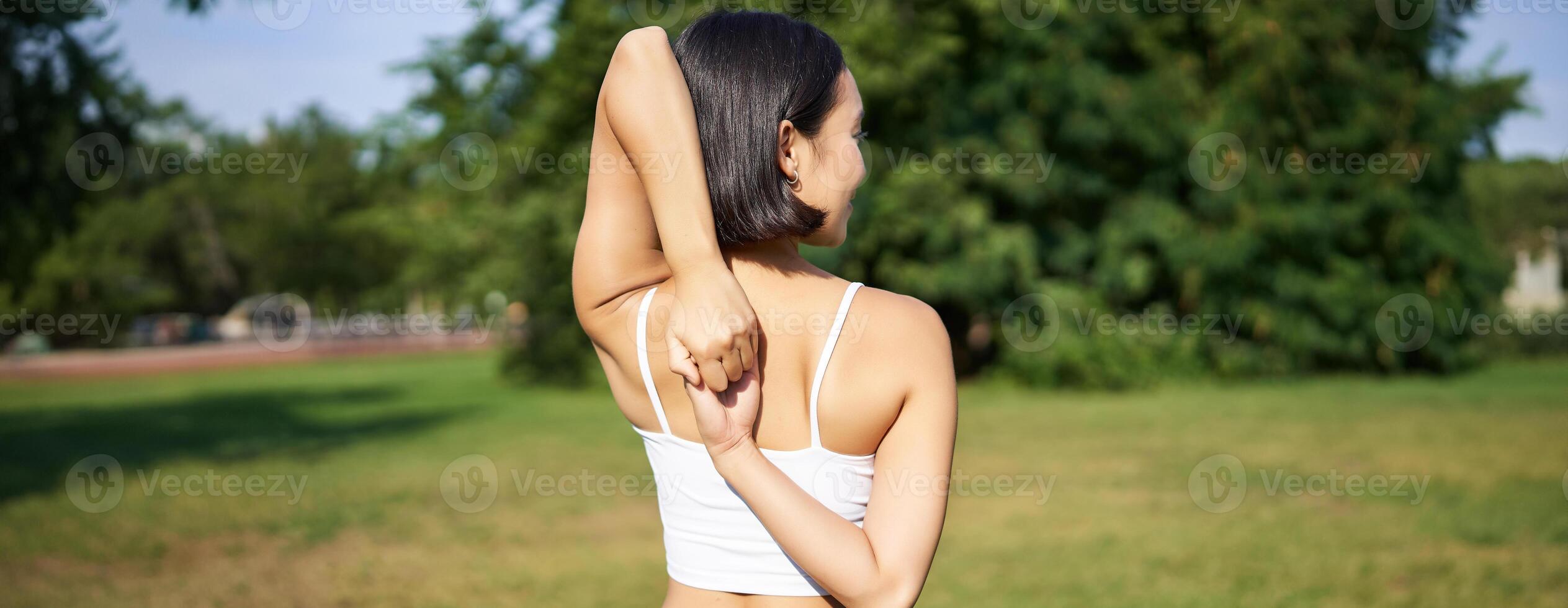 Rear view of young sporty woman stretching her arms behind back, warm-up, prepare for workout jogging, sport event in park photo
