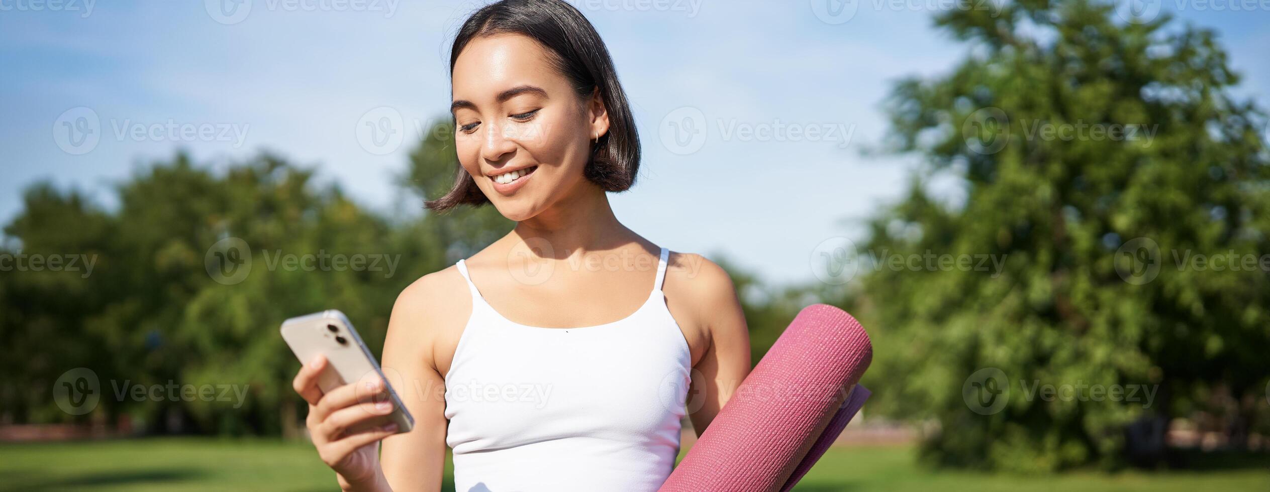 retrato de sonriente asiático mujer con yoga estera, mirando a su teléfono inteligente y leyendo en solicitud, en pie en parque vistiendo deporte uniforme foto