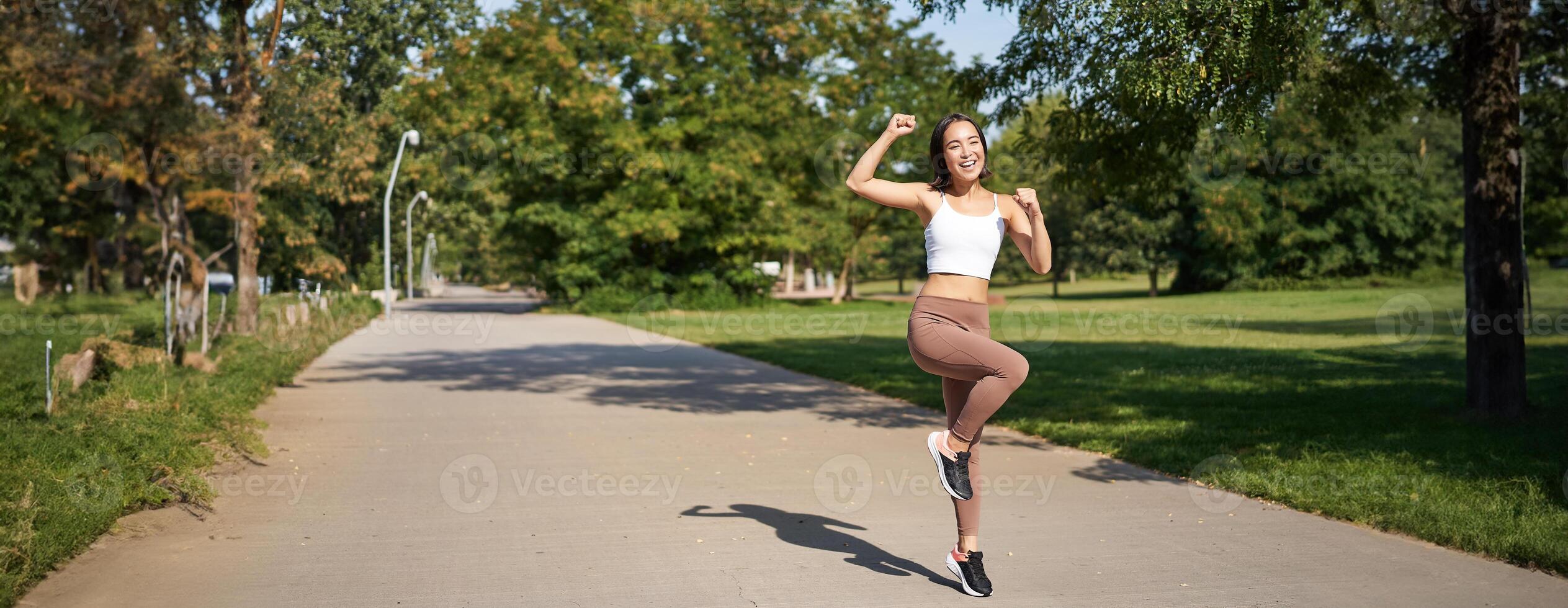 Excited young asian woman winning, finish running in park, saying yes, lifting hand up in triumph, celebrating victory or success photo