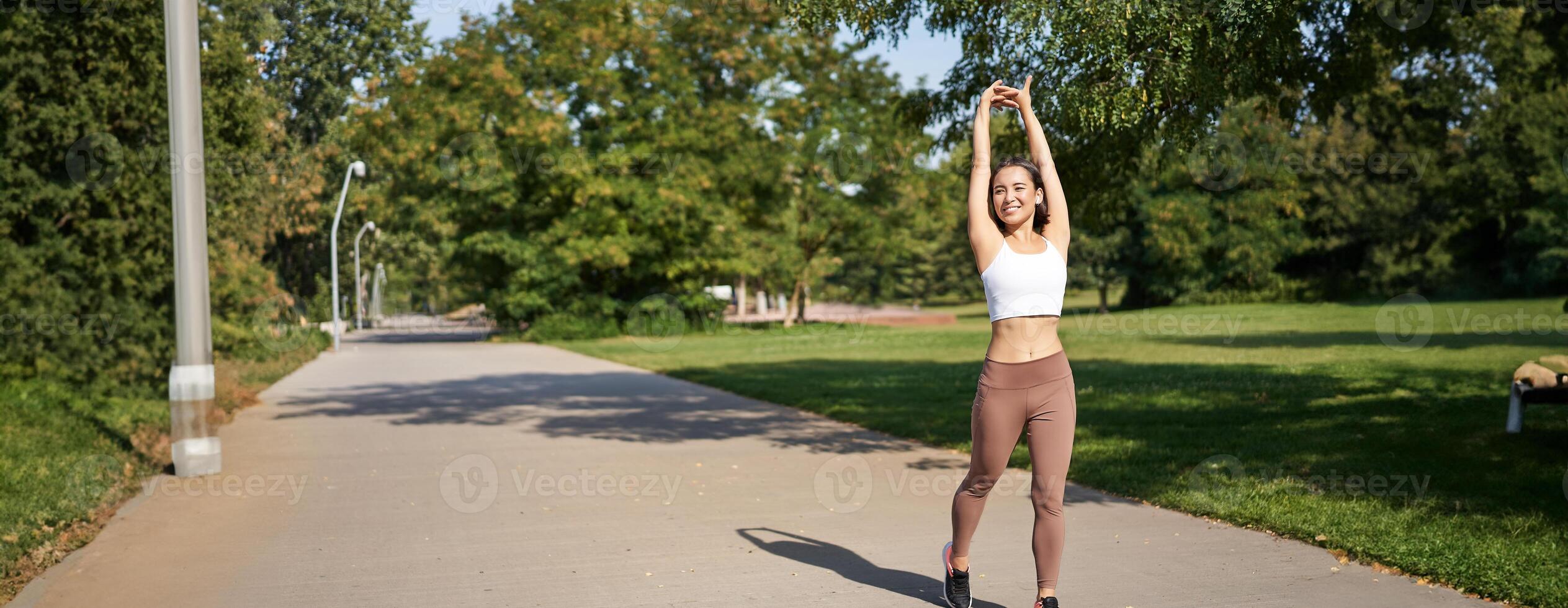 Smiling Asian girl stretching after good workout in park, listening music in wireless headphones, jogging outdoors photo